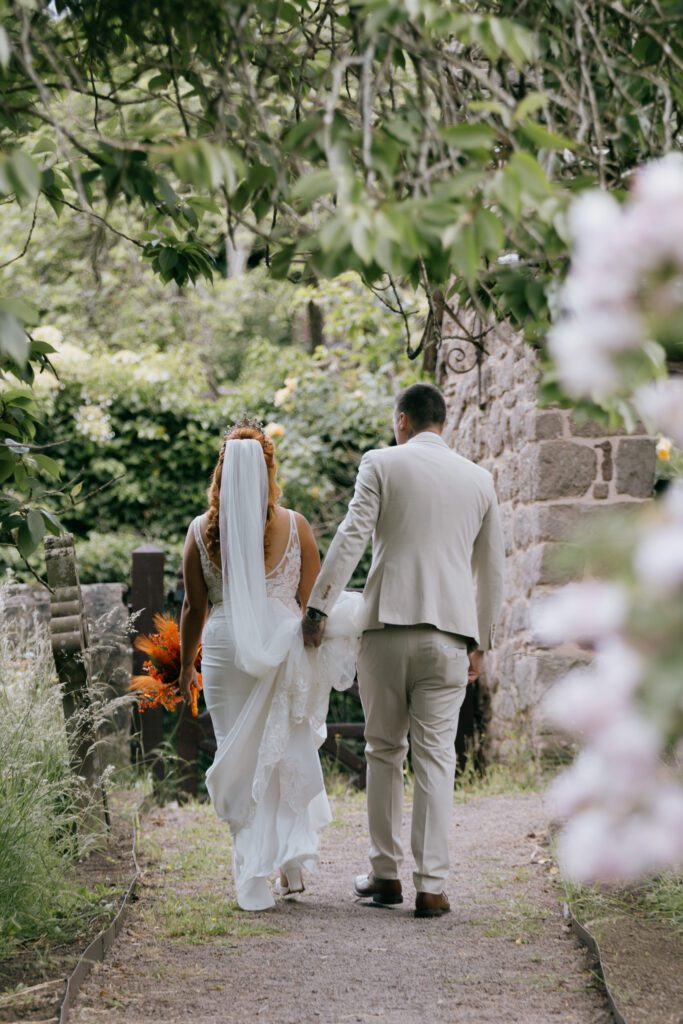 A bride in a white wedding dress and veil holding a bouquet, walking hand-in-hand with her groom in a beige suit along a gravel path surrounded by greenery and stone walls.