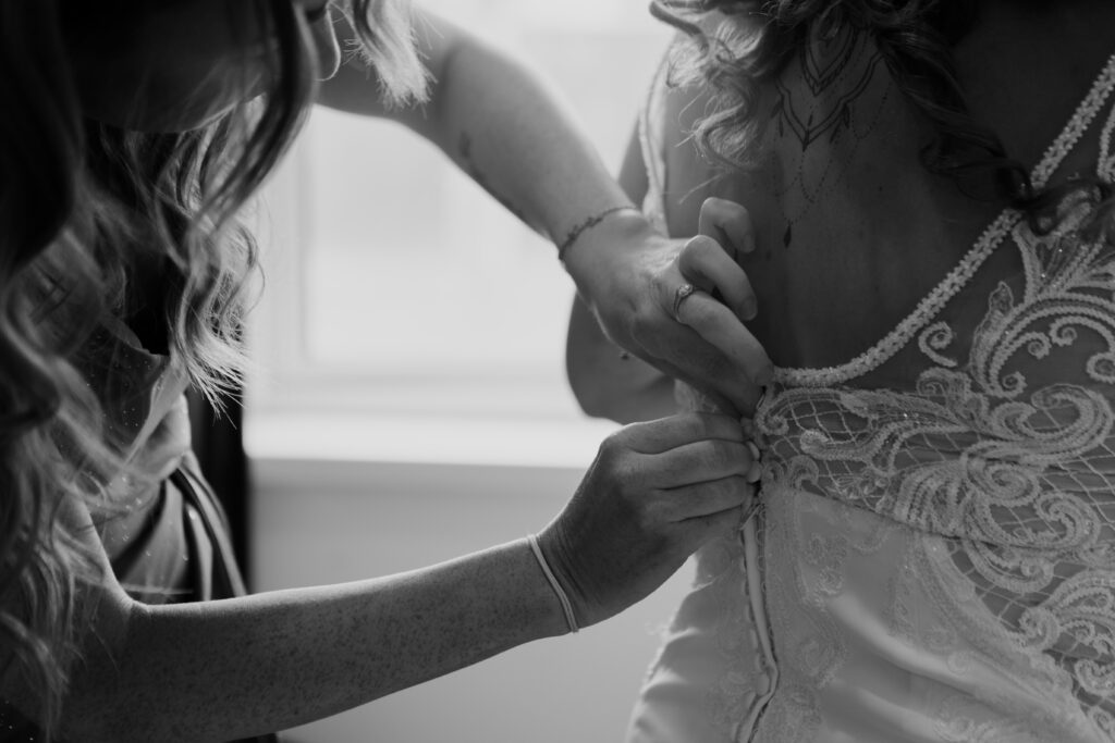 A woman helping another woman fasten the back of a lace wedding dress.
