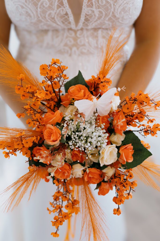 A stunning bridal bouquet featuring orange roses, white roses, and delicate floral accents, complemented by feathery orange embellishments and a white butterfly.