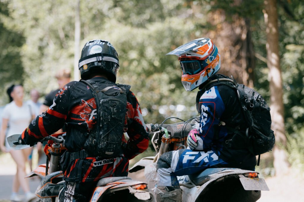 ALT_TEXT: Two motocross riders in gear chat in a wooded area.  

CAPTION_TEXT: Two motocross enthusiasts, fully geared up with helmets and b