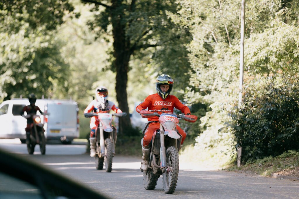 ALT_TEXT: Two motorcyclists in red gear ride dirt bikes on a shaded road.

CAPTION_TEXT: On a sun-dappled road lined by lush greenery, two m