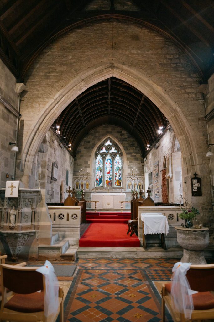 A historic stone church with arched ceilings, a red carpet leading to the altar, and a vibrant stained glass window at the far end.