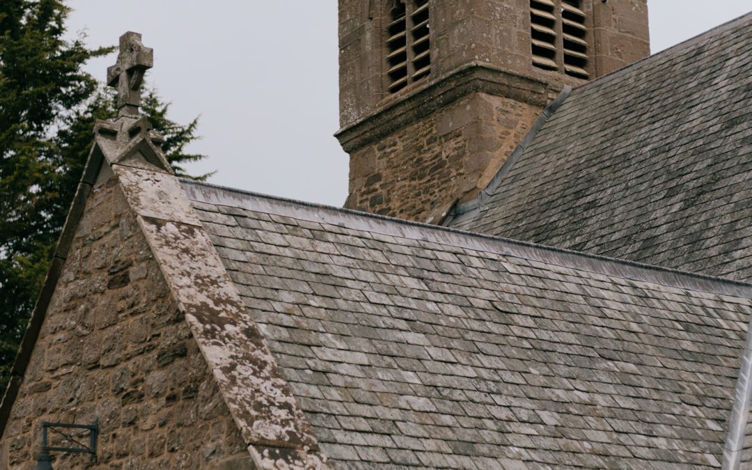 A historic stone church featuring a tall tower with arched windows and a slate roof, set against a cloudy sky.