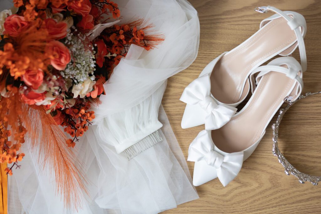 A bridal bouquet featuring orange flowers alongside a pair of white high-heeled shoes with bows, a white veil, and a silver tiara on a wooden surface.