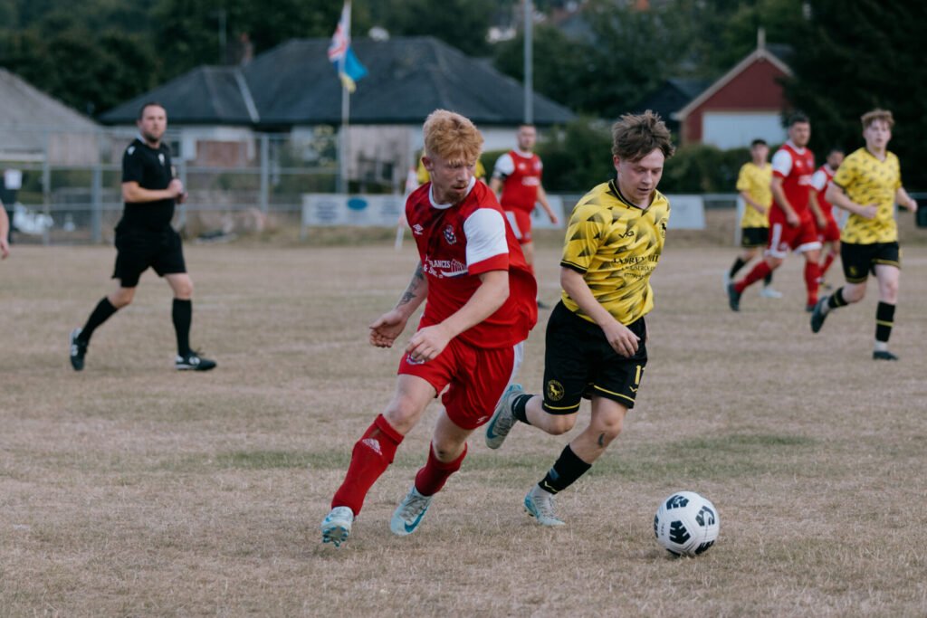 Two football players competing for the ball on a grassy field during a match. One player in a red kit and another in a yellow and black striped kit. A referee and other players are in the background.