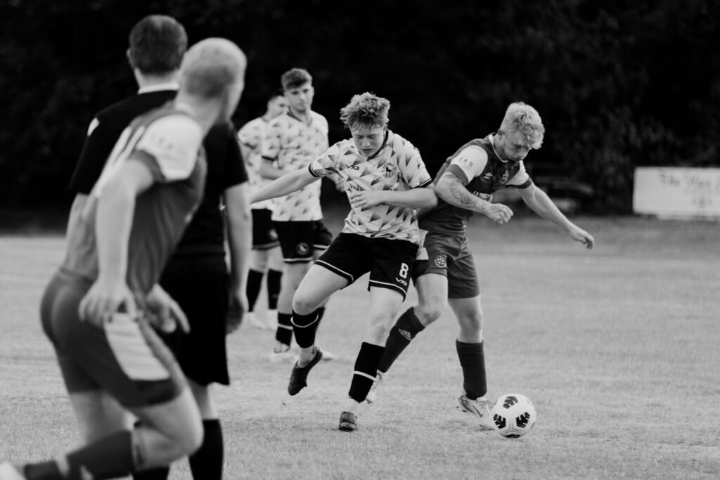Two football players competing for the ball during a match, with several other players visible in the background. One player is in a white jersey with geometric patterns, while the other wears a red jersey. The scene captures the dynamic movement and intensity of the game.