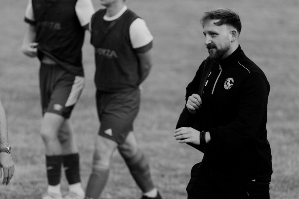 A coach in a black tracksuit with a logo, smiling while walking on a grass football pitch. In the background, players are wearing training vests.