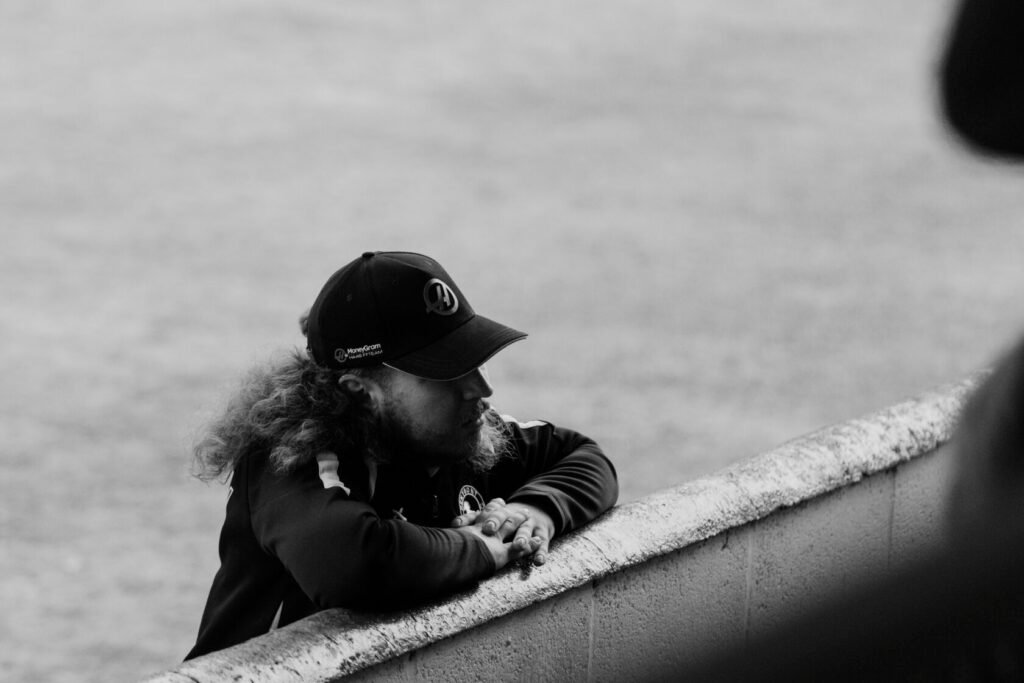 A young man with long curly hair and a cap leans against a concrete barrier, looking contemplative.