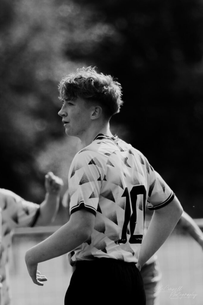 A young football player wearing a patterned jersey with the number 10 stands with a focused expression during a match, captured in black and white.