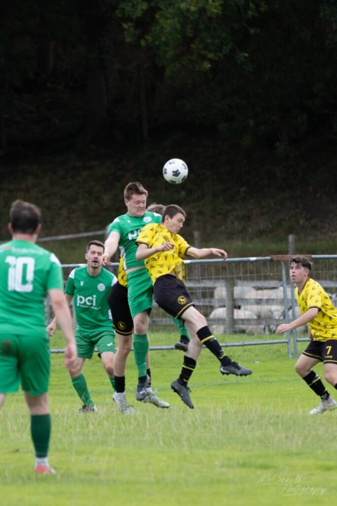 A moment in a football match where players in green and yellow jerseys compete for a high ball. A player in green is attempting to head the ball while another player in yellow leaps to contest it, with additional players in the background.
