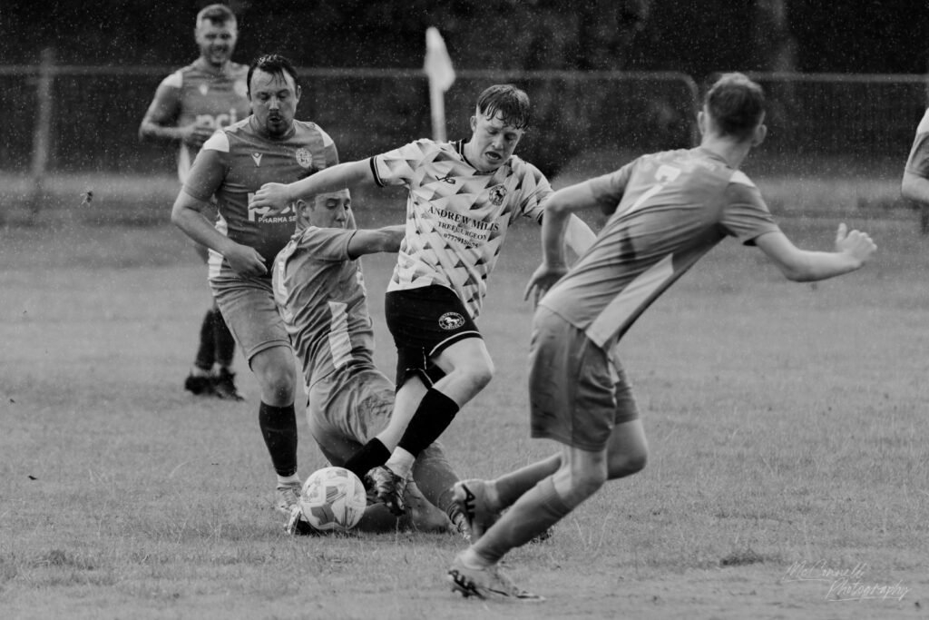 A black and white image of a football match, featuring players engaged in a tackle on a wet field. Water droplets are visible in the air, adding to the intensity of the scene.