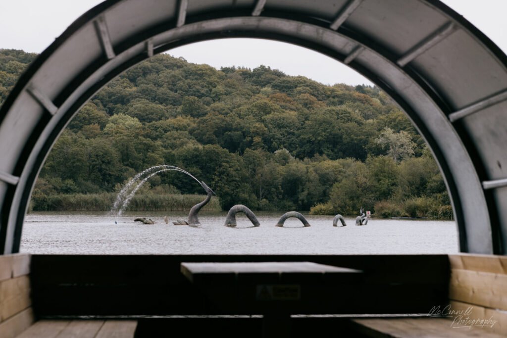 View of a lake with abstract sculptures resembling mythical creatures, seen through a circular arch of a wooden structure.