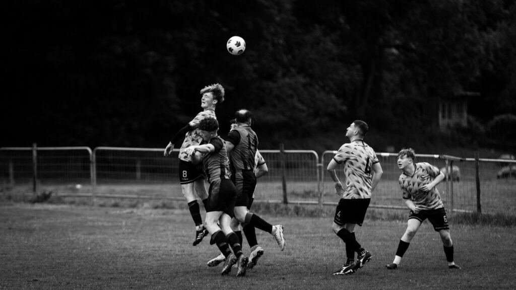 A black and white photograph capturing a group of young football players competing for the ball in mid-air during a match. One player is leaping while others are in close proximity, showcasing dynamic movement and teamwork.