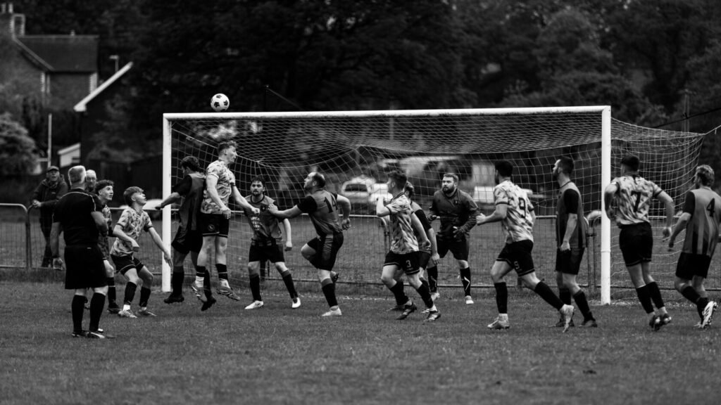 A black and white photograph of a football match with players in various jerseys competing near the goal. A player jumps to head the ball while others react, and spectators can be seen in the background.