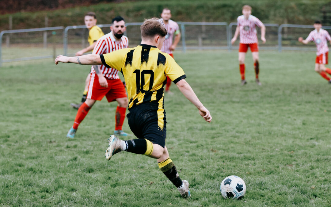 A young football player in a yellow and black striped jersey, wearing number 10, is taking a shot at the ball on a grassy pitch. In the background, players in red and white striped jerseys are positioned nearby.