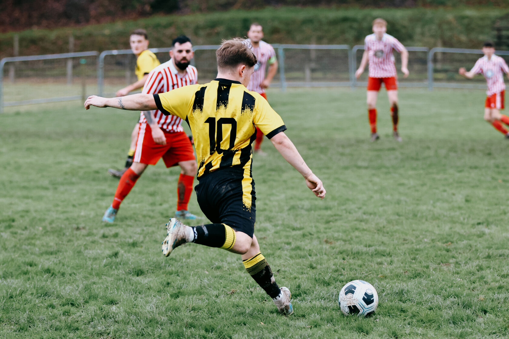 A young football player in a yellow and black striped jersey, wearing number 10, is taking a shot at the ball on a grassy pitch. In the background, players in red and white striped jerseys are positioned nearby.