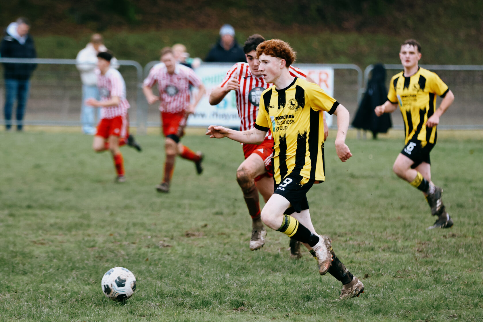 A young football player in a black and yellow striped kit runs with the ball on a muddy pitch, while players in red and white striped shirts chase behind him.
