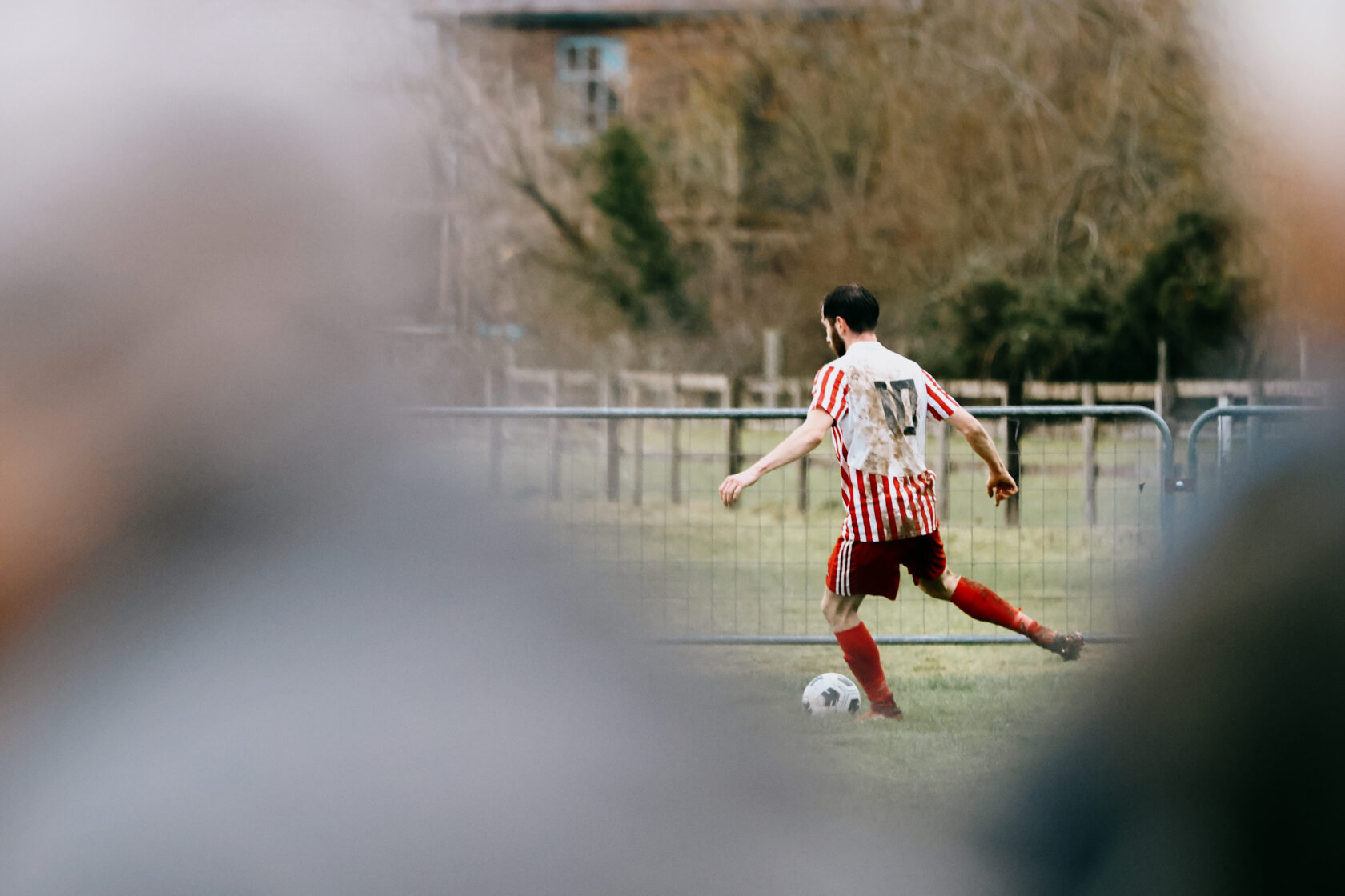 Football Player in Striped Jersey Kicking Ball A football player wearing a red and white striped kit, number 17, kicking a ball on a grassy pitch. The player appears muddy, indicating a competitive match.