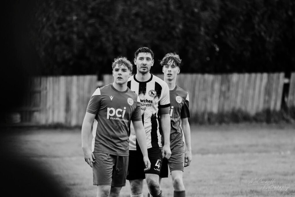 Three football players standing on the pitch, two in grey kits and one in a striped black and white kit, looking focused and serious.