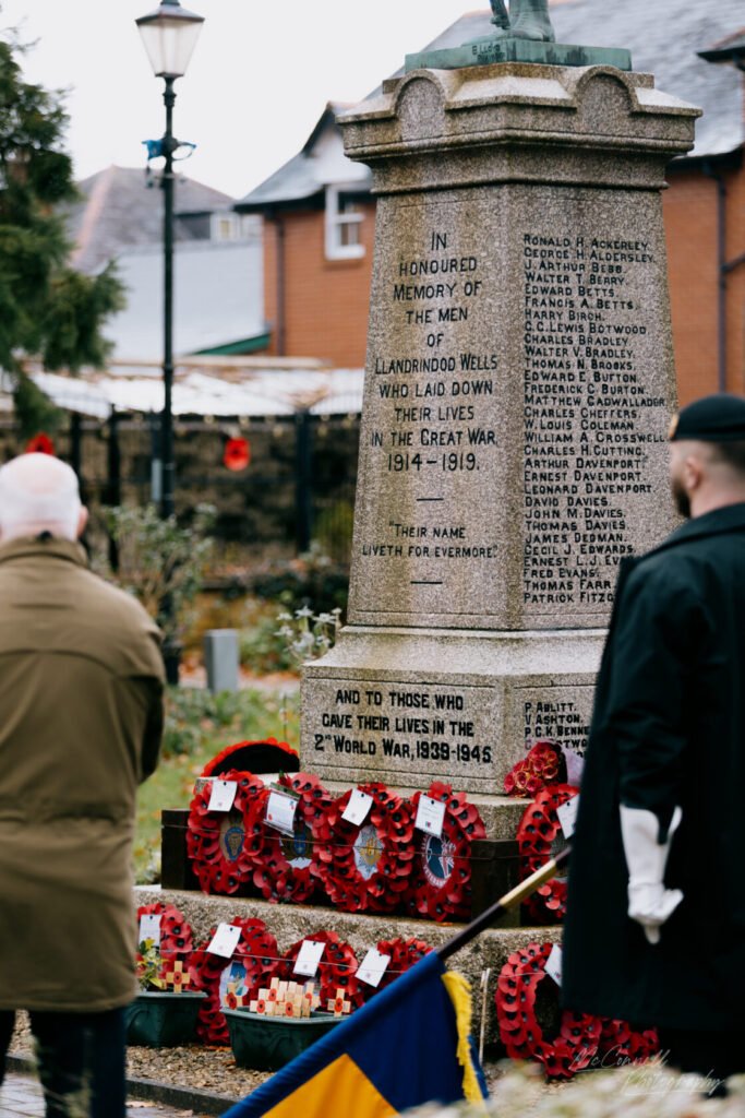 A stone war memorial in Llandrindod Wells inscribed with names of fallen soldiers from the Great War and World War II, surrounded by red poppy wreaths. Two individuals can be seen paying their respects.