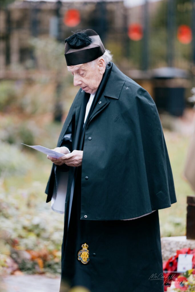 An elderly man dressed in a formal black cape and hat stands outdoors, holding a piece of paper and looking down thoughtfully, surrounded by a natural setting.