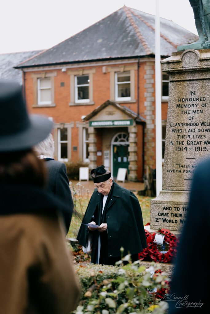 An elderly man dressed in a black coat and hat is reading a document near a war memorial surrounded by autumn foliage and a historical building in the background.
