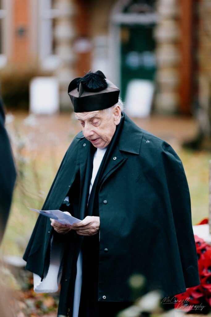 An elderly man dressed in a black cloak and hat reads from a piece of paper during an outdoor ceremony, with a blurred background of autumn foliage and memorial wreaths.