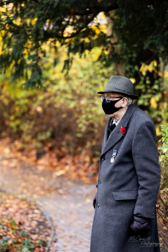 An elderly man wearing a grey coat, a patterned hat, and a black mask stands outdoors surrounded by autumn foliage. He has a poppy pin and medals on his coat, with a pathway visible in the background.