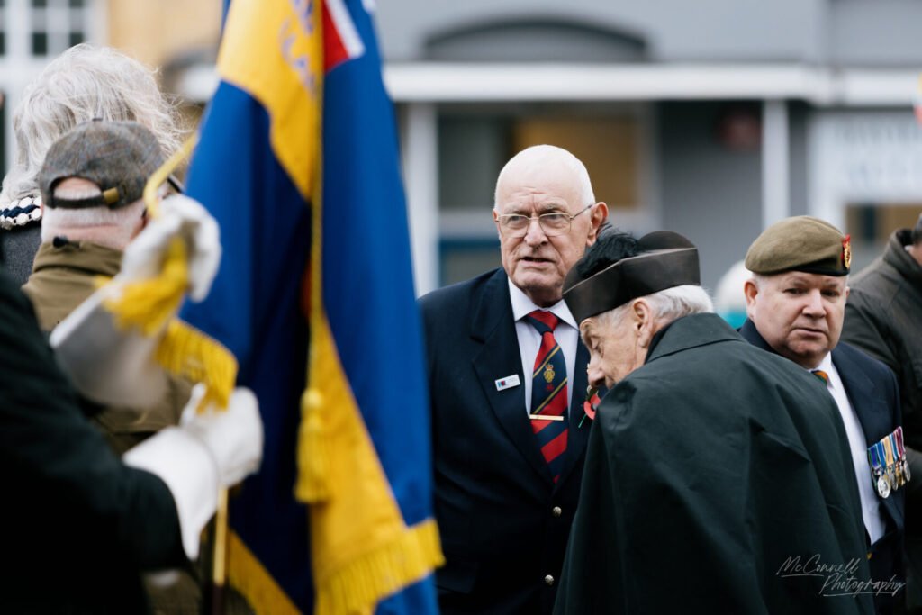 A group of veterans at a remembrance event, with one veteran in a suit and glasses looking intently at another veteran wearing a black cape and a peaked cap, surrounded by flags and military attire.