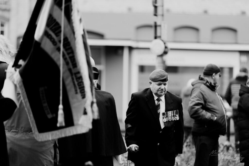 A solemn parade scene featuring a veteran in a formal suit and medals, walking with a cane. Flags, including one for the British Legion, are visible in the foreground with people gathered in the background.