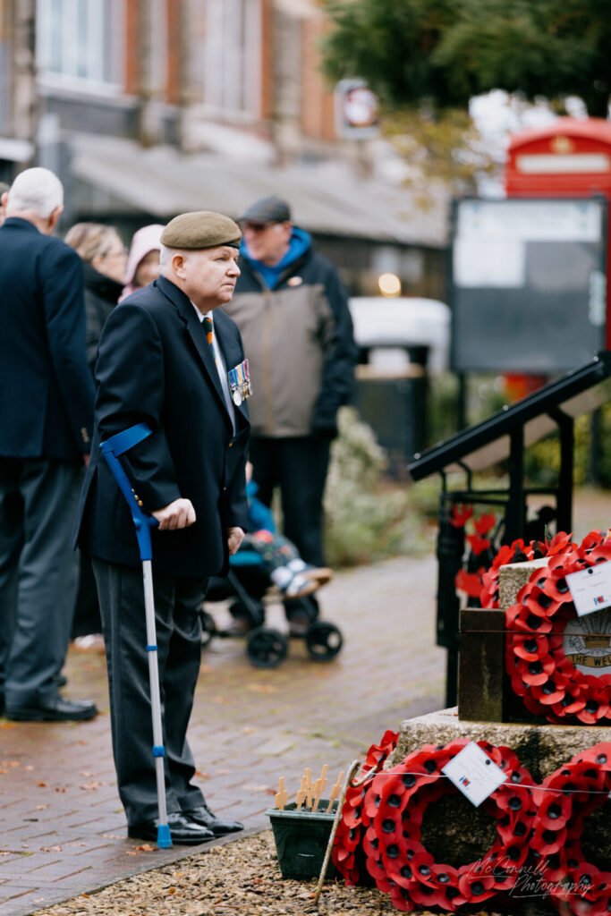 An elderly veteran in military attire, wearing medals and a beret, stands with a cane near memorial wreaths at a remembrance event. The background features people and traditional red telephone boxes.