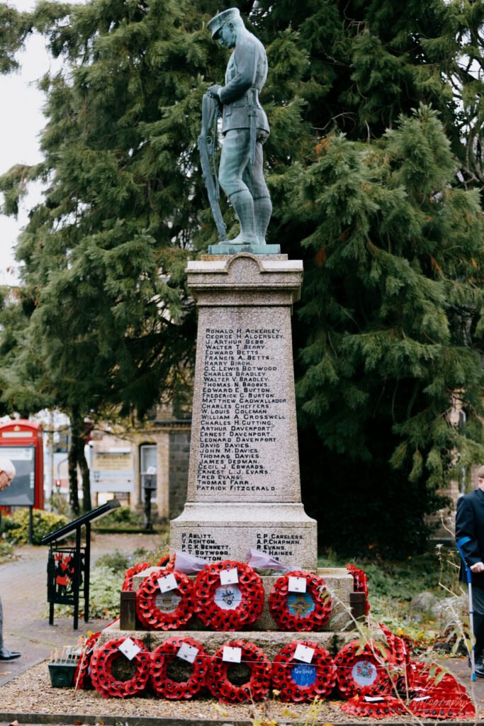 A war memorial statue depicting a soldier, surrounded by red poppy wreaths on the ground. The statue stands on a stone pedestal inscribed with names, set against green trees.