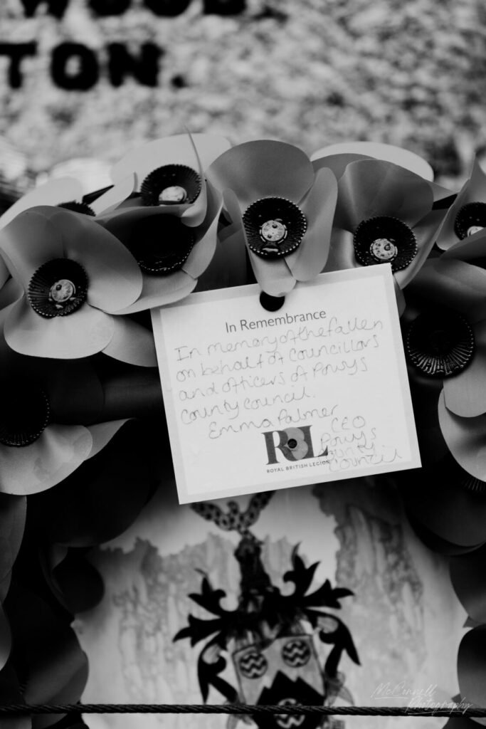 A black and white image featuring a remembrance wreath adorned with black and grey paper poppies. A card is attached, reading 'In Remembrance' with a handwritten message commemorating fallen councillors and officers of a county council.