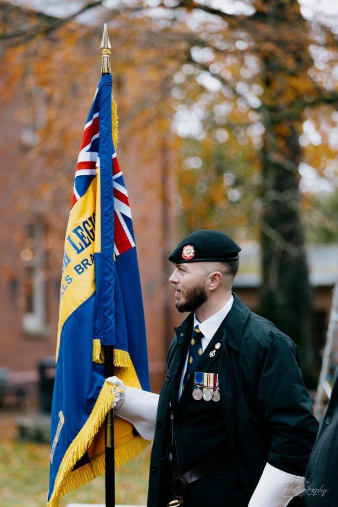 A man in military attire holding a Commonwealth flag, featuring a blue field with a Union Jack and yellow detailing, with autumn foliage in the background.