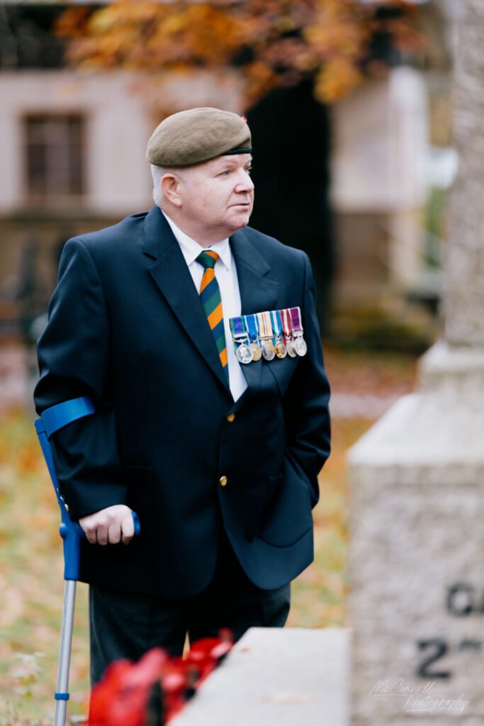 An older man wearing a blazer and beret, adorned with military medals, stands with a blue crutch beside a gravestone, during a remembrance event in an autumnal setting.