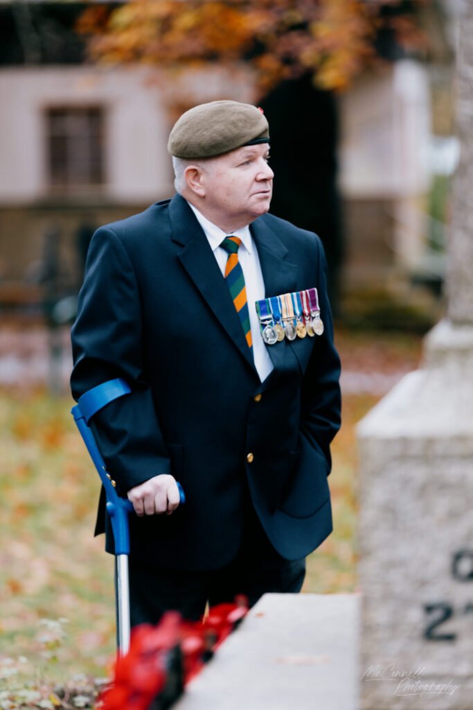 A veteran wearing a dark suit, tie, and military beret stands solemnly with a cane near a gravestone, adorned with medals on his jacket. The background features autumn leaves.