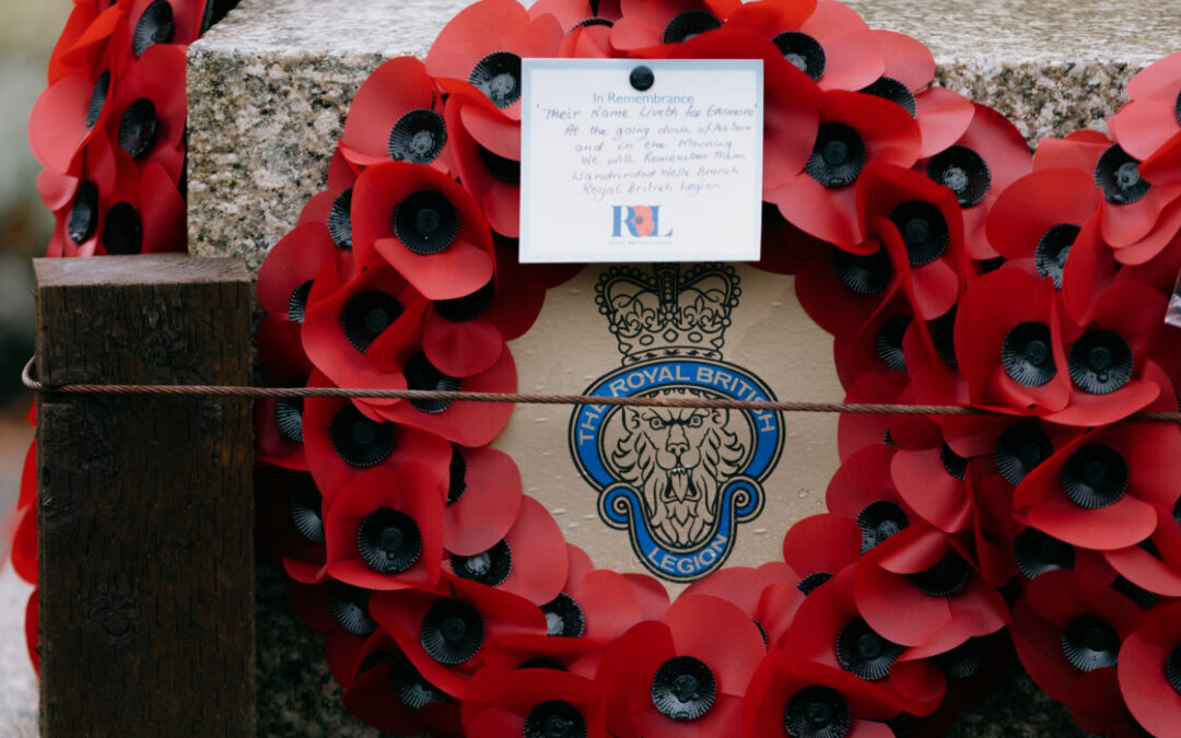 A close-up of memorial wreaths made of red poppies arranged around a stone monument, featuring a card in remembrance. The wreath displays the insignia of the Royal British Legion.