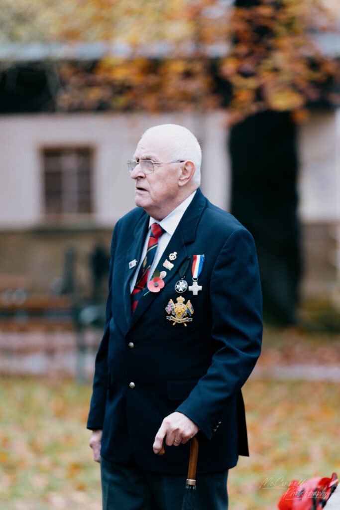 An elderly man wearing a navy blazer adorned with various medals and poppies, standing outdoors with autumn foliage in the background, holding a cane.