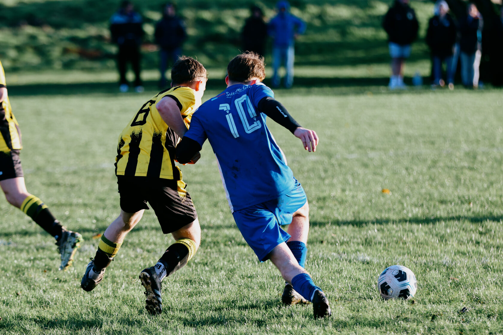 Two young football players competing for the ball on a grassy field, one in a black and yellow striped jersey and the other in a blue jersey with the number 10.