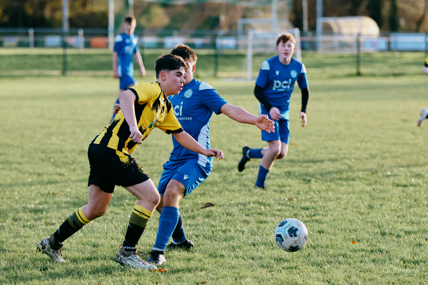A dynamic scene from a youth football match with players in yellow and black jerseys competing against those in blue jerseys for the ball on a green pitch.