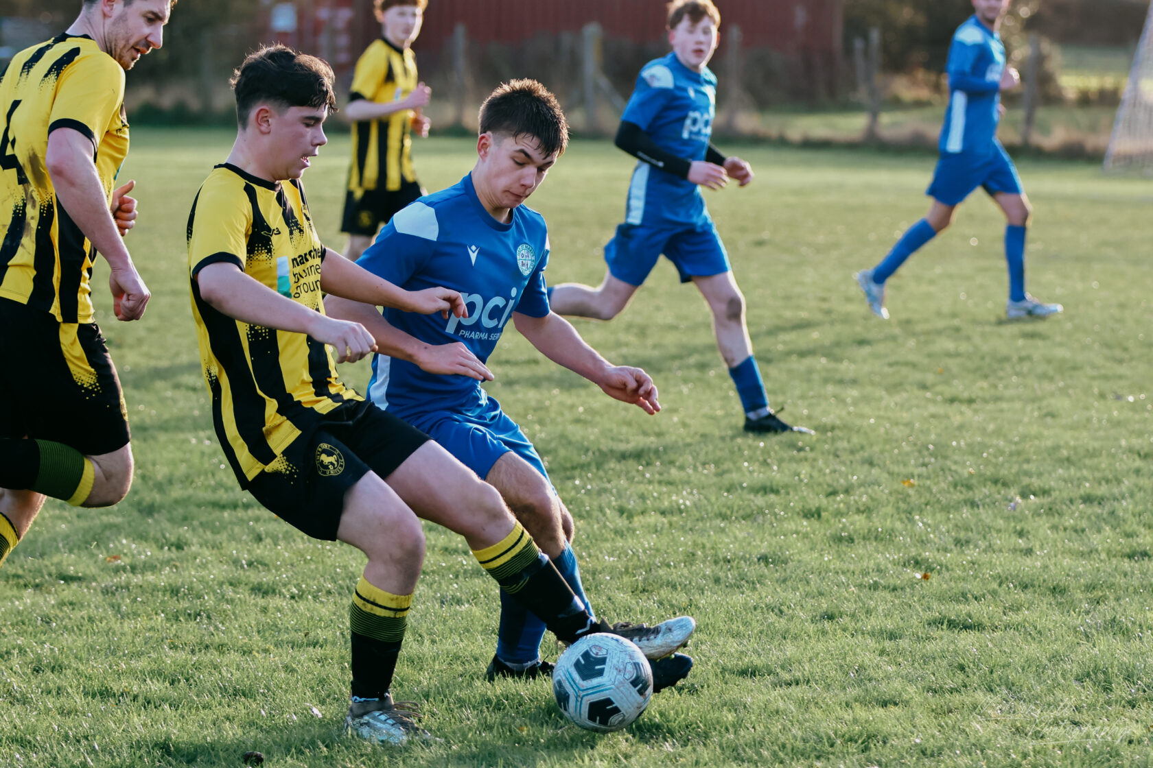 Two football players competing for the ball, one in a blue kit and the other in a black and yellow striped kit, with several other players in the background on a grassy pitch.