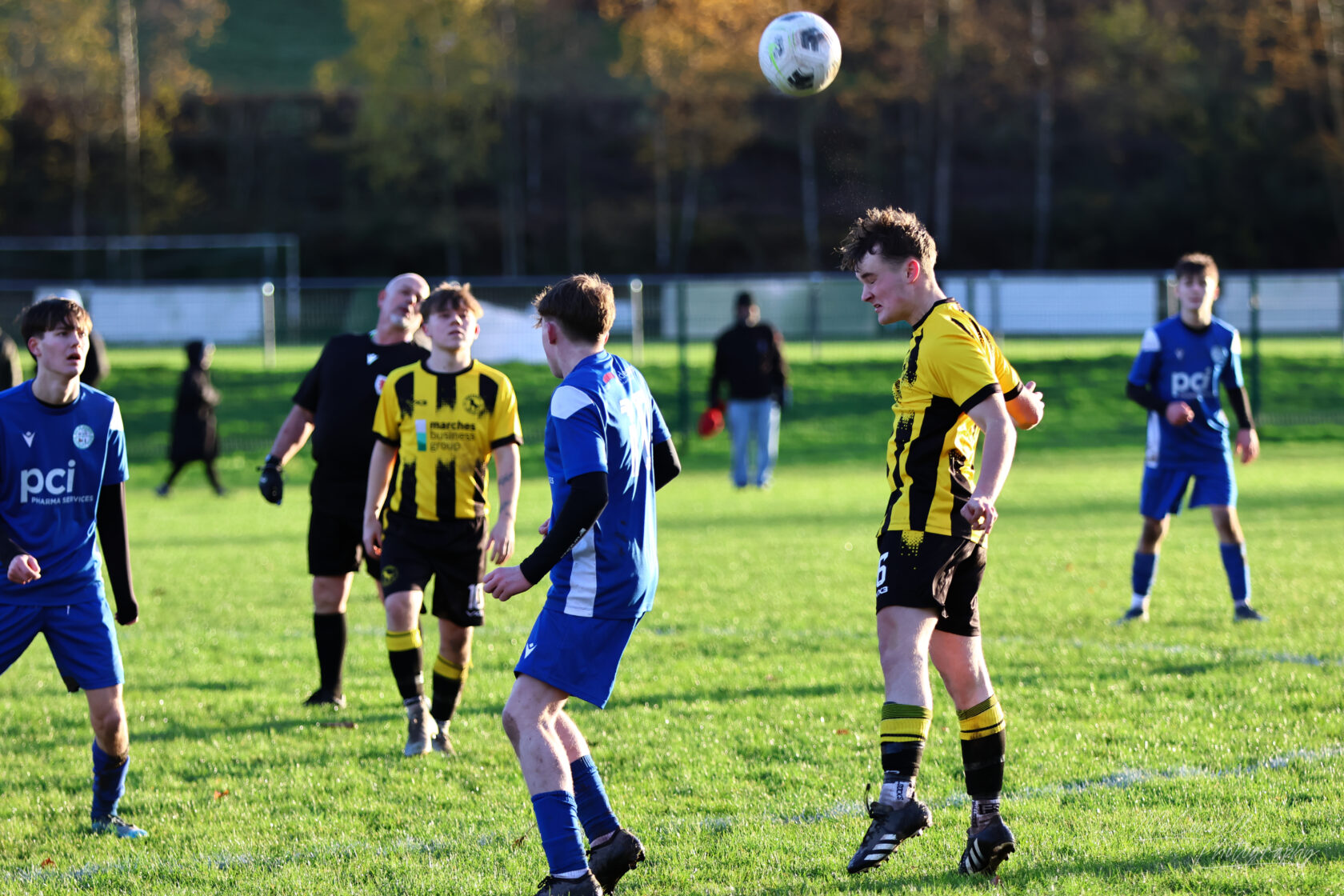 A youth football match in action, featuring players in blue and yellow kits focused on the ball. One player is jumping to head the ball while others are watching. A referee is also present in the background.