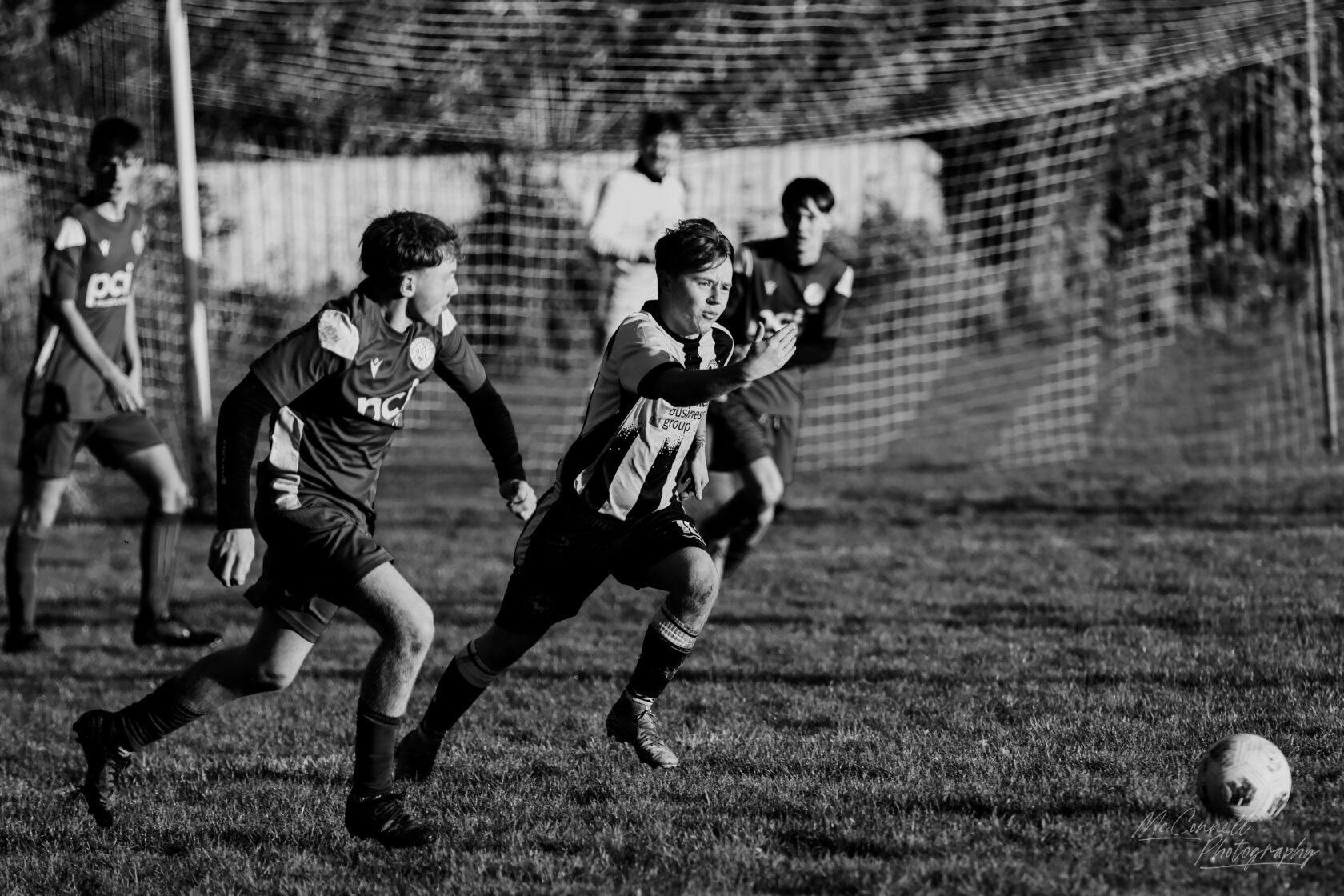 A black and white photograph of a youth football match, featuring two players in motion; one in a striped jersey and the other in a solid coloured jersey, with a goal post in the background and several other players in the scene.