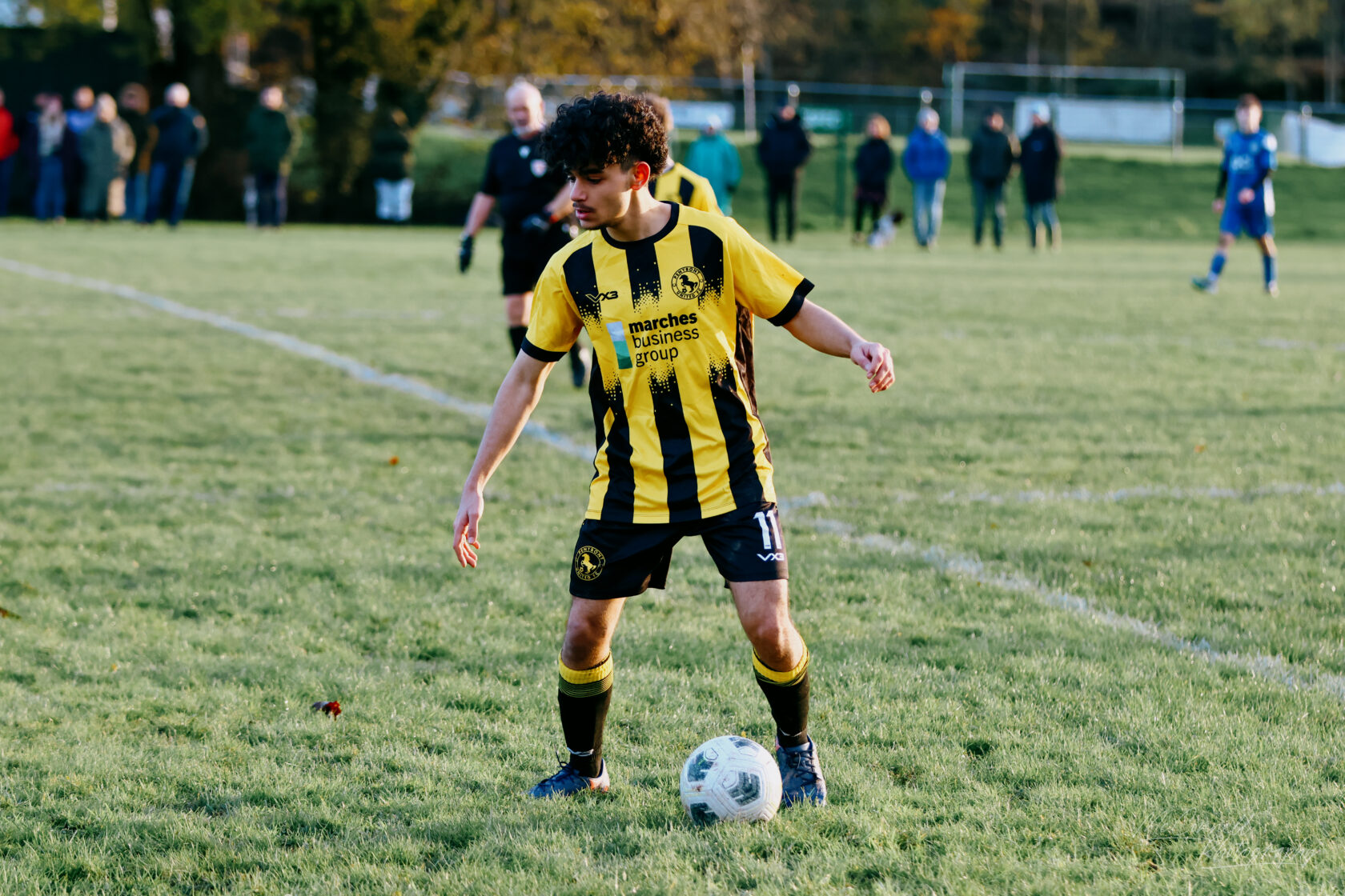 A young male football player wearing a black and yellow striped jersey is poised to kick a football on a grassy pitch, with spectators in the background.