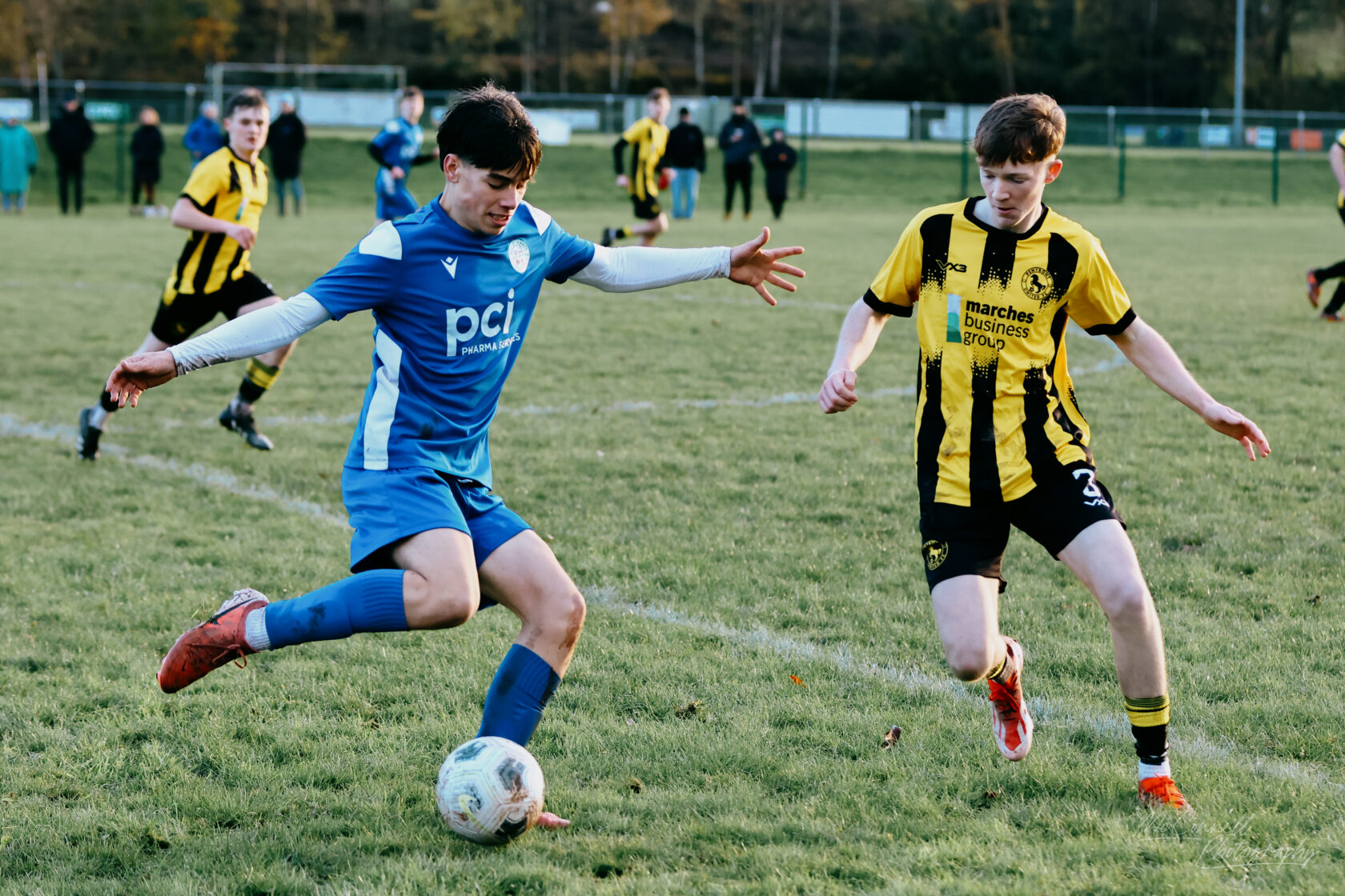 A youth football match in progress with two players competing for the ball. One player in a blue kit is dribbling the ball while a player in a yellow and black striped kit is trying to defend.