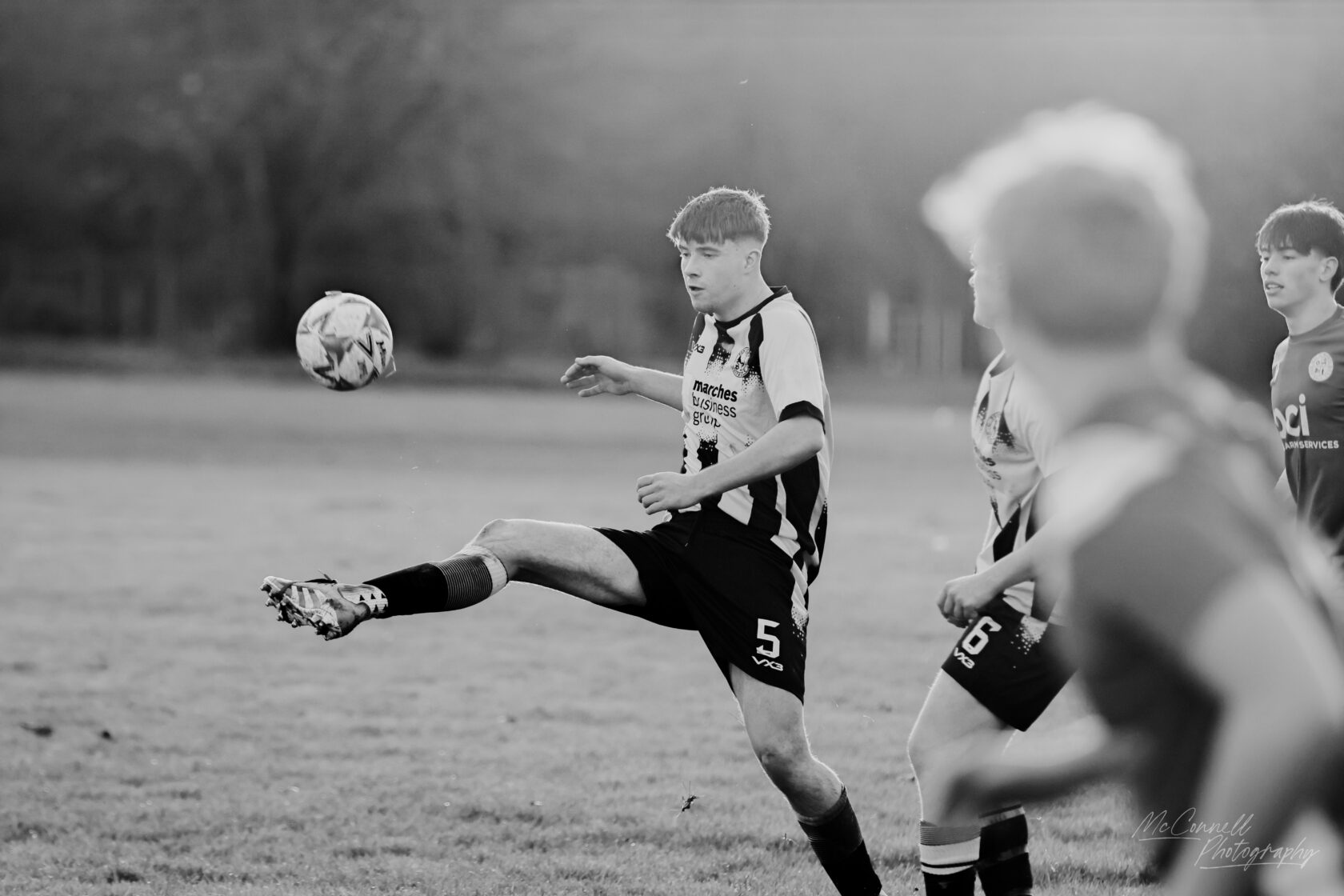 A young football player in a black and white striped jersey kicks a ball during a match on a grassy field, with other players in the background.