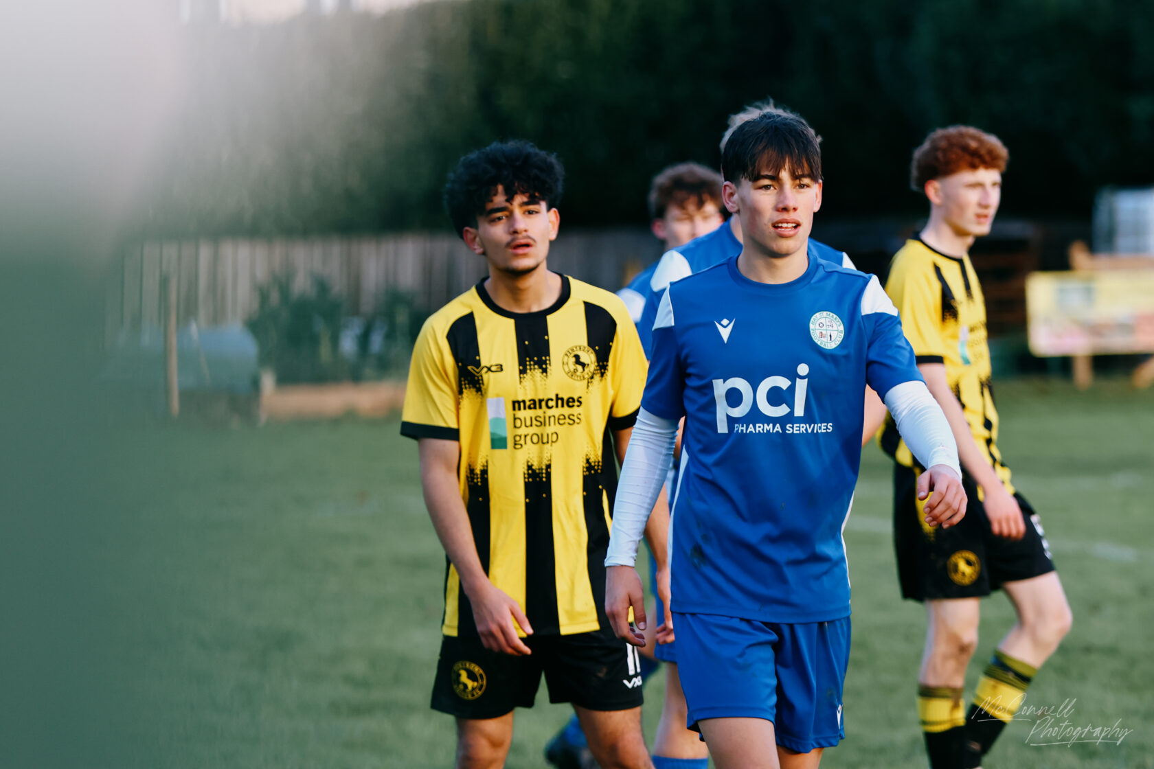 A group of young football players on the field. One player in a blue kit is walking with a focused expression, while a teammate in a yellow and black striped kit stands nearby, looking towards the game.