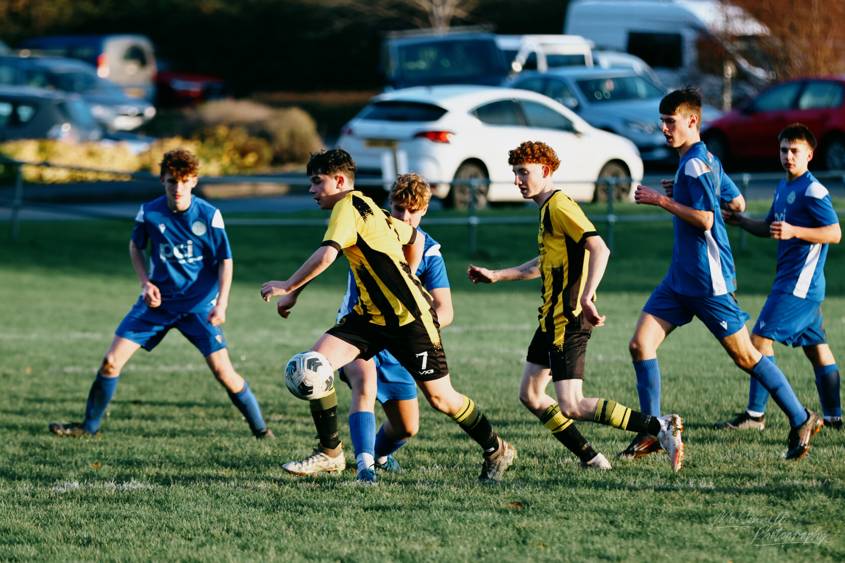 A youth football match in progress, showing players wearing yellow and black striped jerseys and blue jerseys competing for the ball on a grassy field.