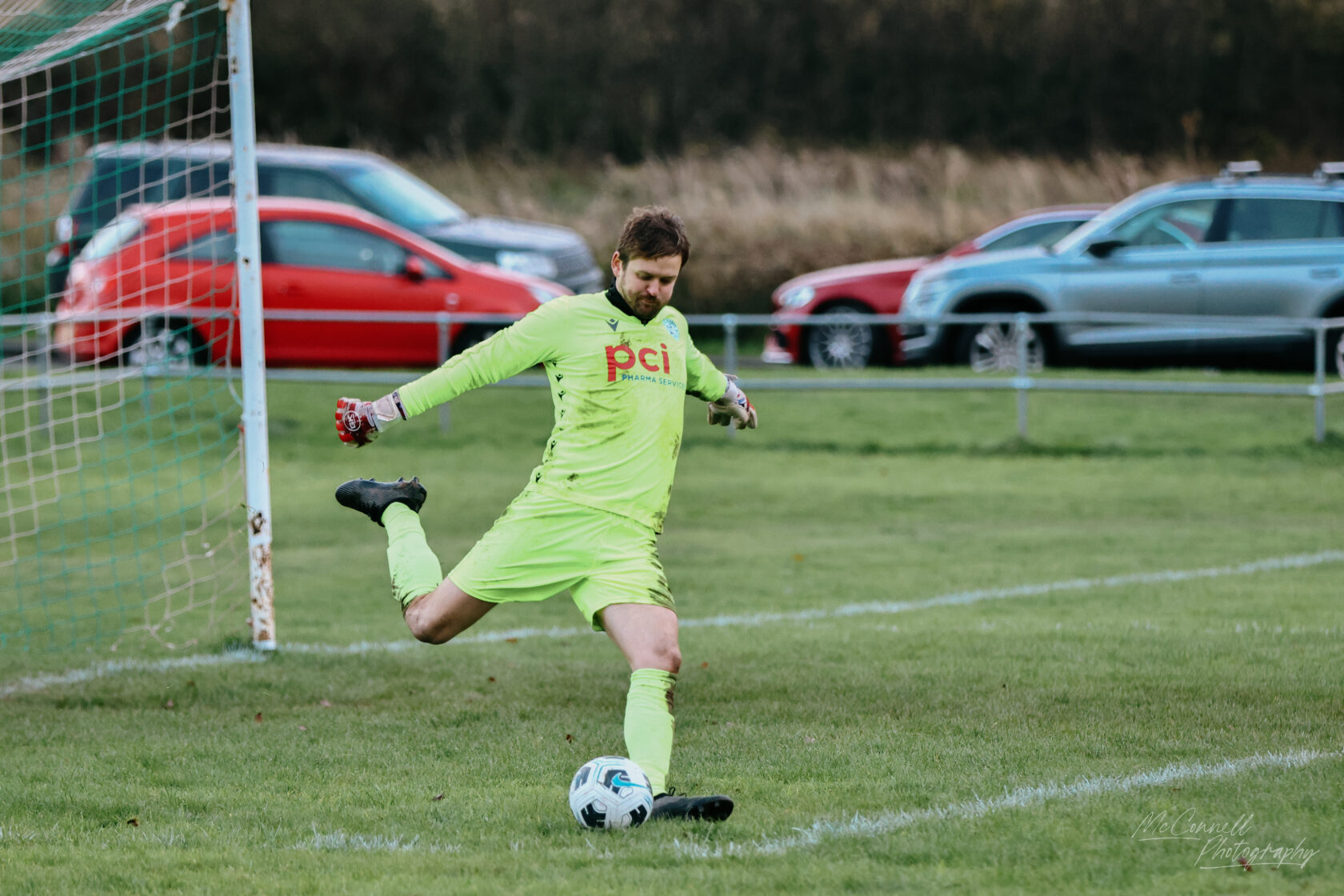 A football goalkeeper in a bright green kit prepares to kick a ball during a match, with a blurred view of cars in the background.