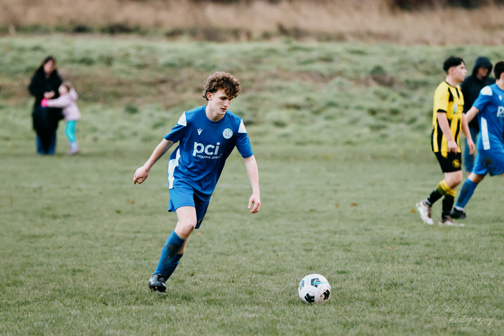 A young football player in a blue jersey dribbles the ball on a grassy field during a youth football match, with other players and spectators in the background.